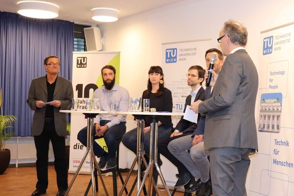 Eröffnung des 26. TU Forums Vize Rektor Fröhlich eröffnet das TU Forum. Hinter ihm sitzt das Podium auf Hochstühlen, links davon steht der Moderator. Im Hintergrund sind TU Rollups zu sehen.