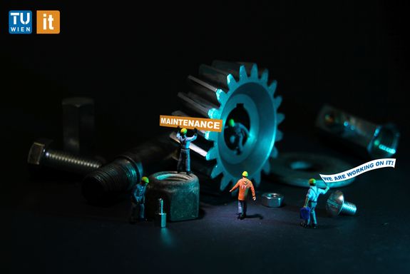 Small figures are surrounded by screws, nuts and gears. The figures wear protective helmets. One figure holds a sign with the inscription "Maintenance", another one carries a flag with the inscription "We are working on it!