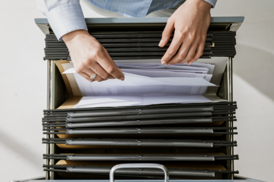 Perspektive von oben: eine Person greift in die Lade eines Aktenschranks und zieht ein Blatt Papier heraus.