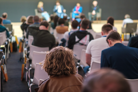 Symbolbild: Das Foto zeigt die sitzenden Zuhörer einer Podiumsdiskussion von hinten und auf der Bühne vier ebenfalls sitzende Personen, die dem Publikum zugewandt, aber auf dem Bild unscharf dargestellt und somit nicht weiter erkennbar sind.