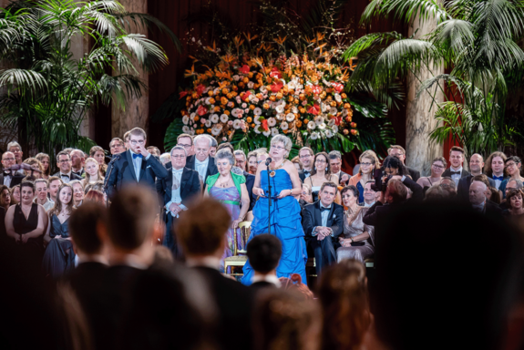Festsaal mit Ballpublikum im Vordergrund, die Rektorin im blauen Ballkleid hält die Eröffnungsrede. Im Hintergrund bunter Blumenschmuck und Palmen.