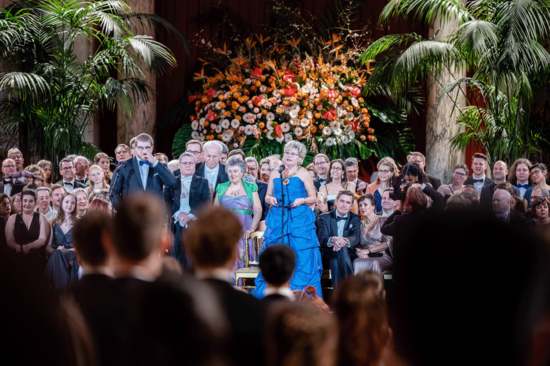 Festsaal mit Ballpublikum im Vordergrund, die Rektorin im blauen Ballkleid hält die Eröffnungsrede. Im Hintergrund bunter Blumenschmuck und Palmen.