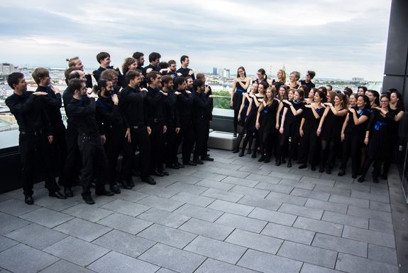 TU Choir There are two groups of people on a roof terrace. On the right is the female group and on the left is the male group. They stand in the same pose (but mirrored) and look at each other.