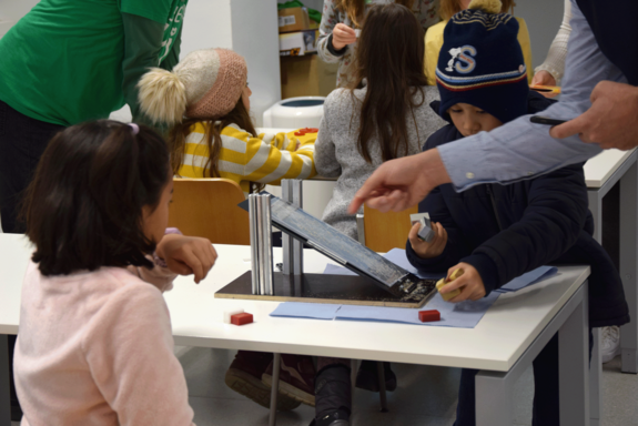 Zwei Kinder an einem Tisch mit einer schiefen Ebene, an der gerade etwas vorgezeigt wird.