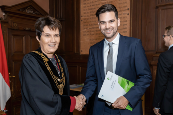 Die Rektorin der BOKU überreicht Preisträger Andreas Stollwitzer seine Urkunde. Sie im Talar, er im dunklen Anzug.