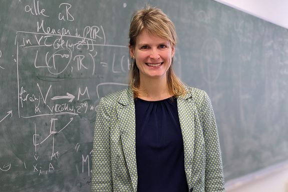 Brustbild. Sandra Müller steht vor einer Tafel beschrieben mit mathematischen Formeln. Sie trägt ihr blondes Haar schulterlang und lächelt. Bekleidung: blaues Oberteil mit grün-weiß gemustertem Jackett.