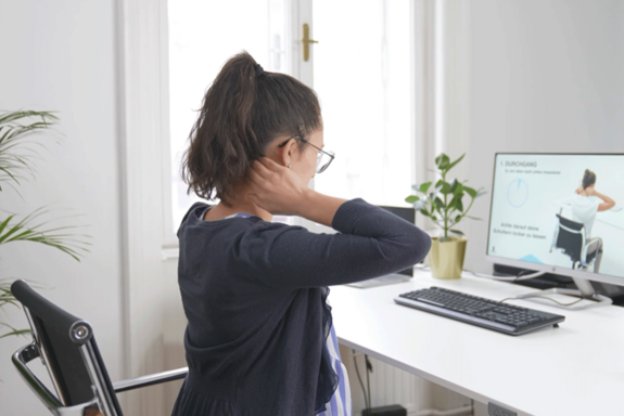 Vor einem Schreibtisch sitzende Person mit einem Pferdeschwanz und Brille hält die Hände am Nacken und macht eine Übung, die ihr am vor ihr stehenden Bildschirm gezeigt wird.