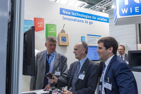 three men infront of the Turbulence Surpression flight simulator, the one in the middle is controlling it – at the TU Wien stand
