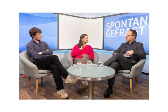 Drei Personen sitzen in einem Fernsehstudio. Vor ihnen ein runder Glastisch mit Wassergläsern. Im Hintergrund weißer Schriftzug auf blauem Hintergrund "SPONTAN GEFRAGT".