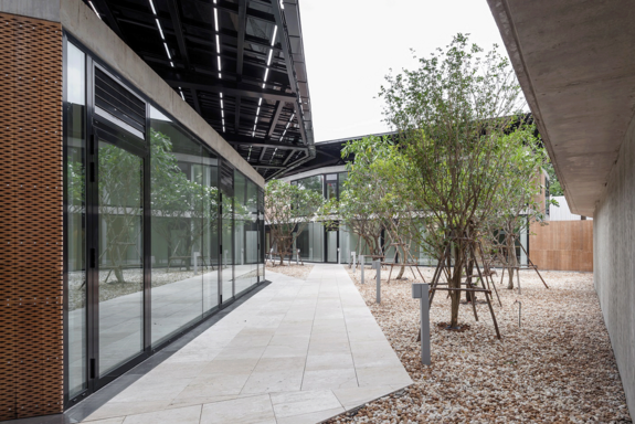 Blick auf den kleinen Innenhof eines modernen Gebäudes mit verglaster Wand, links zu sehen. Rechts von der Wand Pflasterung, wieder rechts davon geschotterte Fläche mit Bäumen.