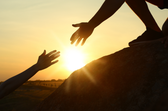A person on a large stone extends a helping hand to another person below. The sunrise can be seen in the background.