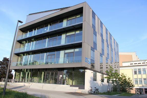Vor blauem Himmel im Vordergrund der Neubau des Atominstituts, dahinter klein zwei Teile des Altbaus.