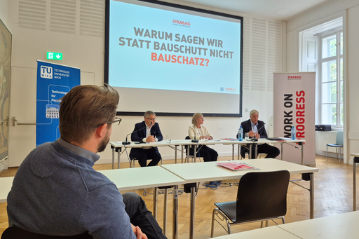 3 Personen sitzend an Tischen als Podium mit Mikrofonen, dahinter Leinwand, links im Bild Mann mit blauem Pullover von hinten