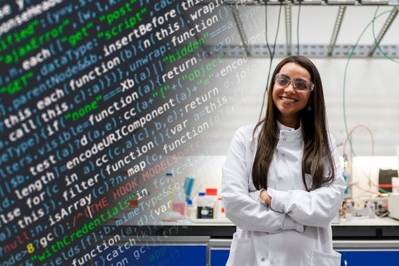 Gut gelaunte Frau mit Schutzbrille und weißem Kittel im Labor, in der linken Hälfte des Bildes ein bunter Computercode der von schräg unten ins Foto läuft.