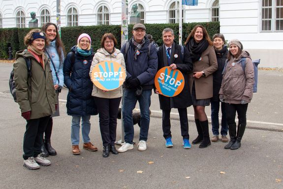 Gruppenbild mit neun Personen. Zwei davon halten große Buttons mit der Aufschrift "Stoppt Gewalt gegen Frauen" in Deutsch und Englisch in Händen.