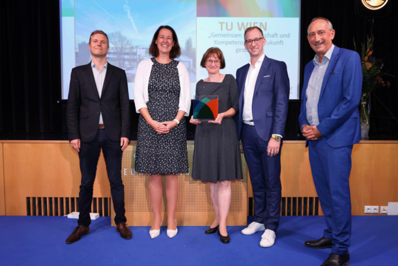 Gruppenfoto mit 5 Personen, 2 in der Mitte mit dunklen Kleidern, links mit dunklem Anzug, links 2 mit dunkelblauen Anzügen. Screen im Hintergrund, blauer Boden.