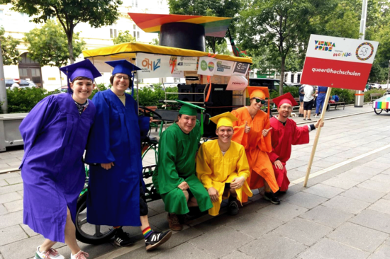 Sechs Menschen in Talaren in den Farben des Regenbogens vor einer bunten Fahrradrikscha.
