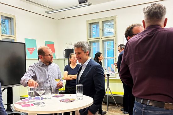 Zwei Männer stehen an einem Stehtisch und unterhalten sich. Weitere Personen von hinten zu sehen.