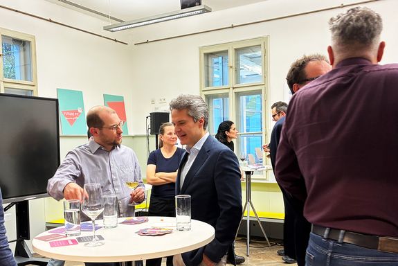 Zwei Männer stehen an einem Stehtisch und unterhalten sich. Weitere Personen von hinten zu sehen.