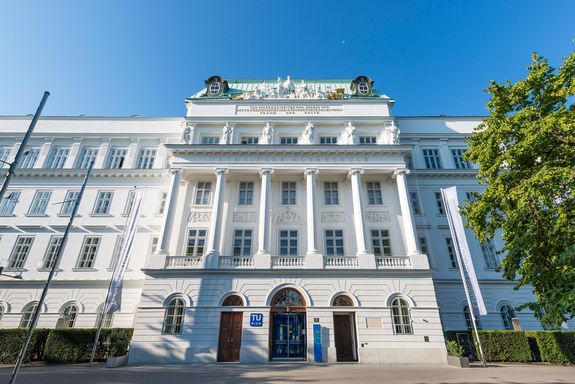 TU Wien Hauptgebäude Frontansicht. Das Gebäude ist länglich, in der Mitte ist der Eingagsbereich mit Säulen und Statuen.