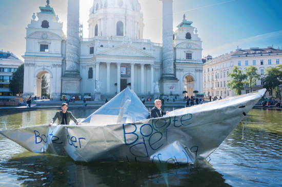 Riesiges Papierfaltboot mit zwei Menschen am Wasser, im Hintergrund die Karlskirche.