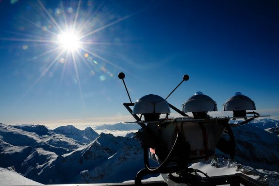 Measuring instruments in front of a wintry mountain backdrop. Above, a cloudless sky and the sun.