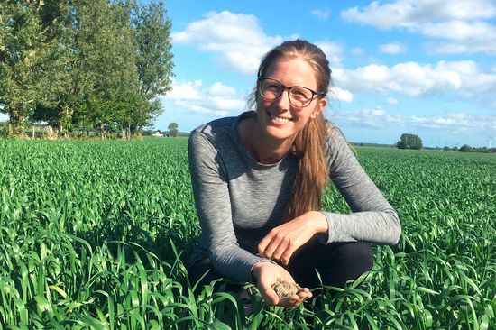 Mariette Vreugdenhil bei blauem Himmel auf einem Feld, im Hintergrund Bäume. In der Hand hält sie ein Häufchen Erde.