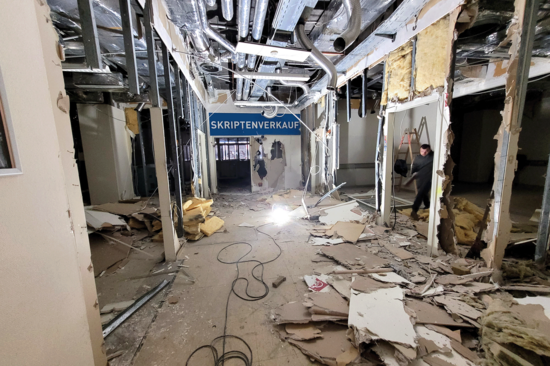 Baustelle Campus Freihaus mit herausgerissenen Wänden und sichtbaren Röhren am Plafond, am Boden liegen herausgerissene Platten. Rechts, ein Arbeiter, am Ende des Ganges ein blaues Schild mit weißer Schrift „SCHRIFTENVERKAUF“.
