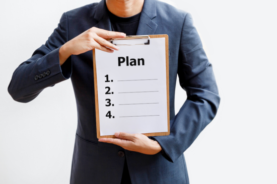 Ein Person mit Blazer hält sich ein Klemmbrett vor die Brust auf dem "Plan" steht.