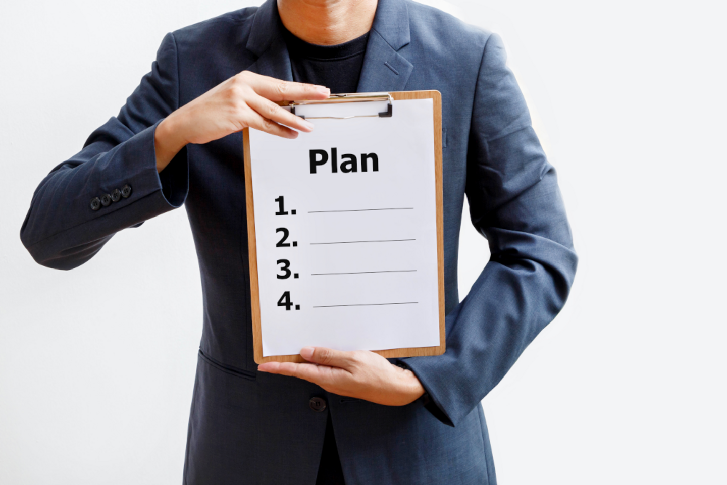 A person in a blazer holds a clipboard in front of his chest with ‘Plan’ written on it.