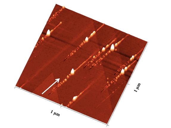 Nan Nanostrukturen nach dem Ionenbeschuss: Der Pfeil zeigt die Richtung der Ionen an: Zunächst ensteht ein Graben mit Nanohillocks an beiden Seiten, ein besonders großer Hügel zeigt den Impakt-Punkt an, danach ist noch eine Erhebung entlang des Pfades zu sehen
