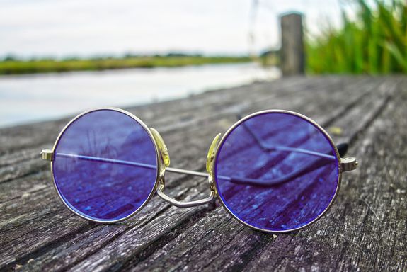 Blue-purple tinted round sunglasses in the foreground lie on a summery wooden walkway.