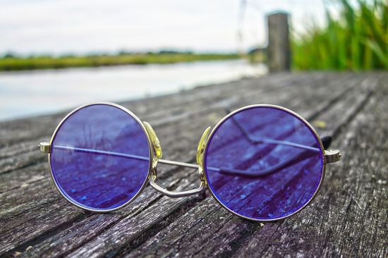 Blue-purple tinted round sunglasses in the foreground lie on a summery wooden walkway.