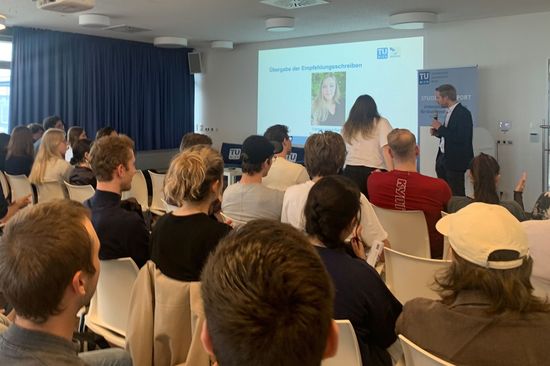 Publikum von hinten sitzend, Vortragender stehend mit Mikro bei Leinwand