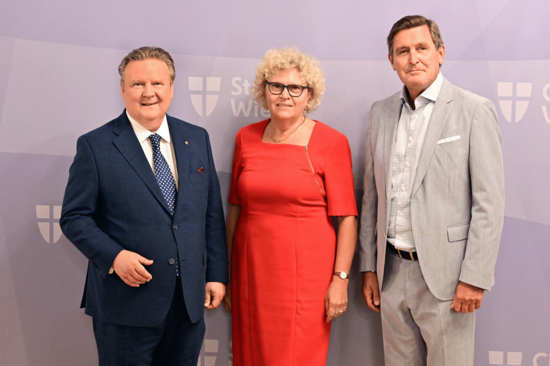 Gruppenfoto mit drei Personen, die ab den Knien aufwärts abgebildet sind. Bürgermeister Ludwig im blauen Anzug, Altrektorin Seidler im roten Kleid, Stadtrat Hanke im hellen Anzug. Hintergrund lila mit Stadt-Wien-Logos. 
