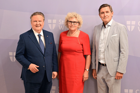 Gruppenfoto mit drei Personen, die ab den Knien aufwärts abgebildet sind. Bürgermeister Ludwig im blauen Anzug, Altrektorin Seidler im roten Kleid, Stadtrat Hanke im hellen Anzug. Hintergrund lila mit Stadt-Wien-Logos. 