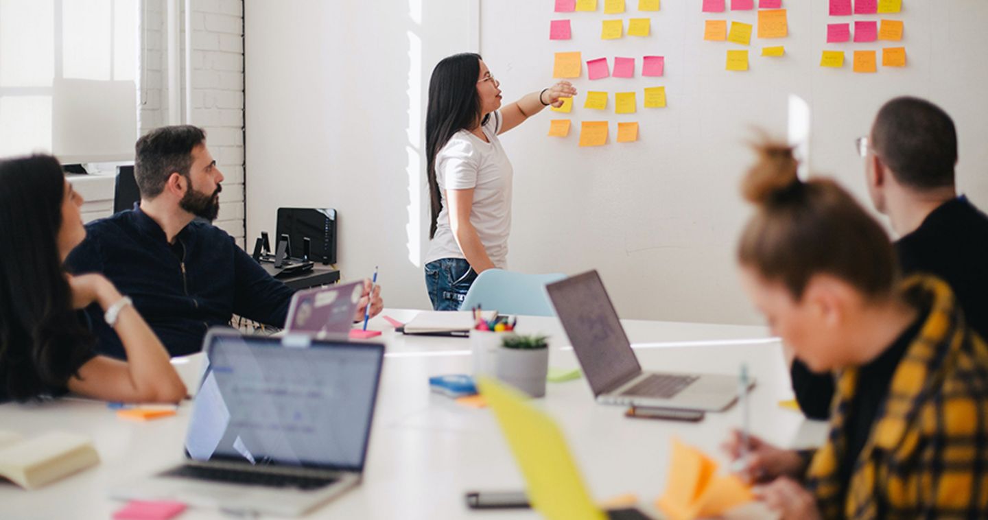 A woman points to colourful sticky notes on the wall and several people sit at a table.
