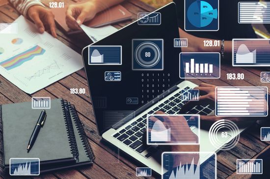 Tisch mit Laptop, Zetteln und Notizbüchern, links oben im Bild Hände, rechts in weiß Diagramme über das Bild gelegt