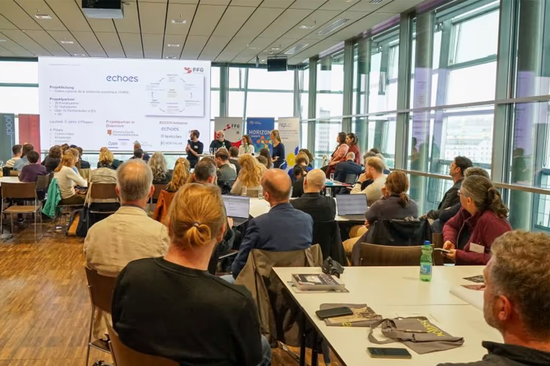 Das Foto zeigt einen großen Raum mit Personen, die gruppiert an Tischen sitzen. Im Hintergrund ist eine Präsentation auf einer Leinwand zu sehen, davor stehen und sitzen vier weitere Personen.