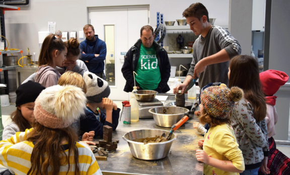Kinder bei ihrem Workshop im Labor an einem Arbeitstisch mit Metallbehältern. Die Kinder im Vordergrund tragen gelb-weiß gestreifte Pullover, eines eine Kappe, eines eine Haube.