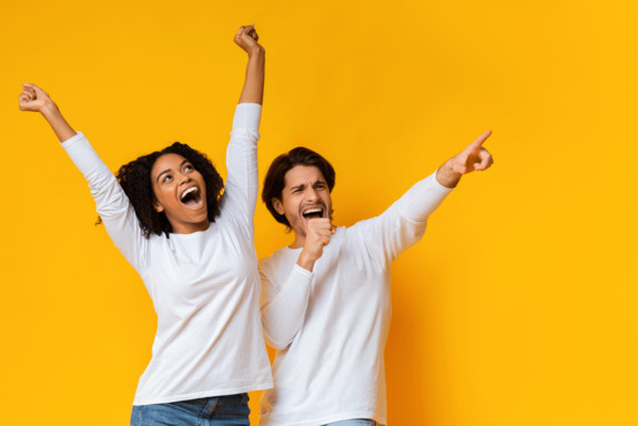 Junges Paar mit weißen Shirts, das die Arme jubelnd in die Höhe streckt. Gelber Hintergrund.