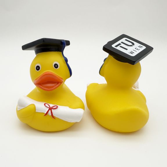 2 Quietscheenten in Gelb, beide tragen einen typischen Absolvent_innen-Hut. Die linke Ente schaut in die Kamera und hält eine Promotionsrolle in Weiß. Die rechte Ente dreht den Rücken zu, sodass man auf dem Hut das TU Wien Logo erkennen kann. 