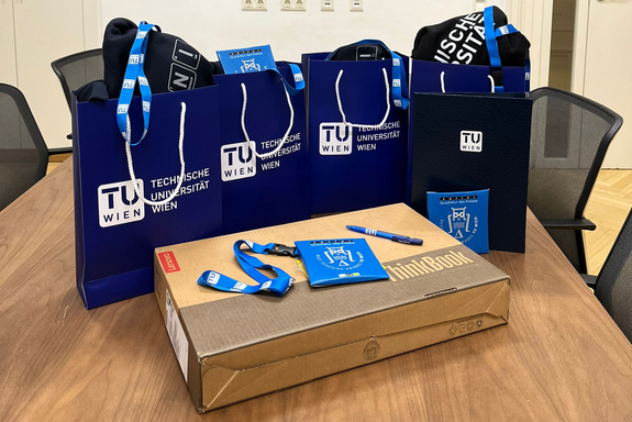 brauner Tisch, darauf braune Laptopschachtel, dahinter blaue Sackerl mit TU Logo