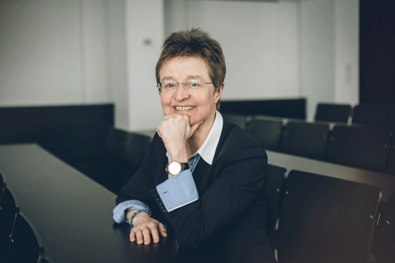 Dekanin Gerti Kappel sitzt lächelnd in einem Seminarraum, eine Hand am Pult abgelegt, mit der anderen stützt sie ihr Kinn. Gekleidet ist sie mit einem blauen Jakett und heller Bluse.