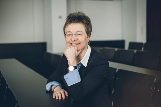 Dekanin Gerti Kappel sitzt lächelnd in einem Seminarraum, eine Hand am Pult abgelegt, mit der anderen stützt sie ihr Kinn. Gekleidet ist sie mit einem blauen Jakett und heller Bluse.