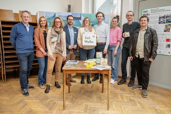 Neun Personen stehen nebeneinander, die Mittlere hält ein Schild mit der Aufschrift "Soil Walks", im Vordergrund ein Tisch.
