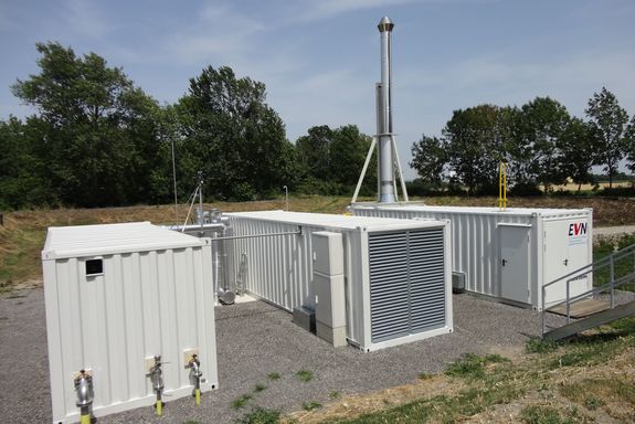 3 Container auf einem Schotterplatz im Grünen – einer mit dem EVN Logo und Zugangstüre, einer mit Lüftungsgitter und Stromkasten und einer mit 3 Anschlüssen, die in die Erde führen. Ziwschen den Containern sind silberen Rohrleitungen zu sehen.