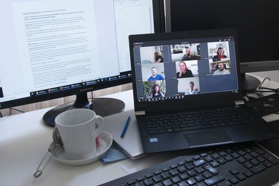 Ein heimischer Schreibtisch mit Kaffeetasse und aufgeklapptem Laptop, auf dem eine Videokonferenz mit 8 teilnehmenden Personen zu sehen ist