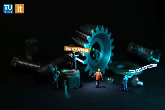 Kleine Figuren sind von Schrauben, Müttern und Zahnrädern umringt. Die Figuren tragen Schutzhelme. Eine Figur hält ein Schild mit der Aufschrift "Wartung", eine andere trägt eine Fahne mit der Aufschrift "Wir arbeiten daran!"
