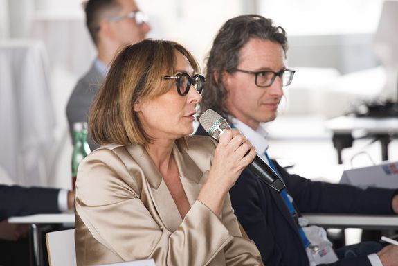 A woman sits and speaks while holding a microphone. Two men are sitting in the background.
